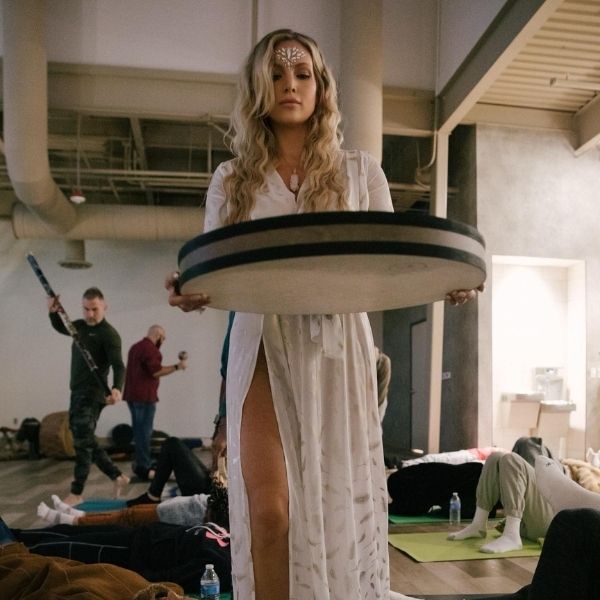 A Healing Sanctuary facilitator leading a sound bath during a Live Community REset Experience, holding a frame drum while participants relax in a meditative state.